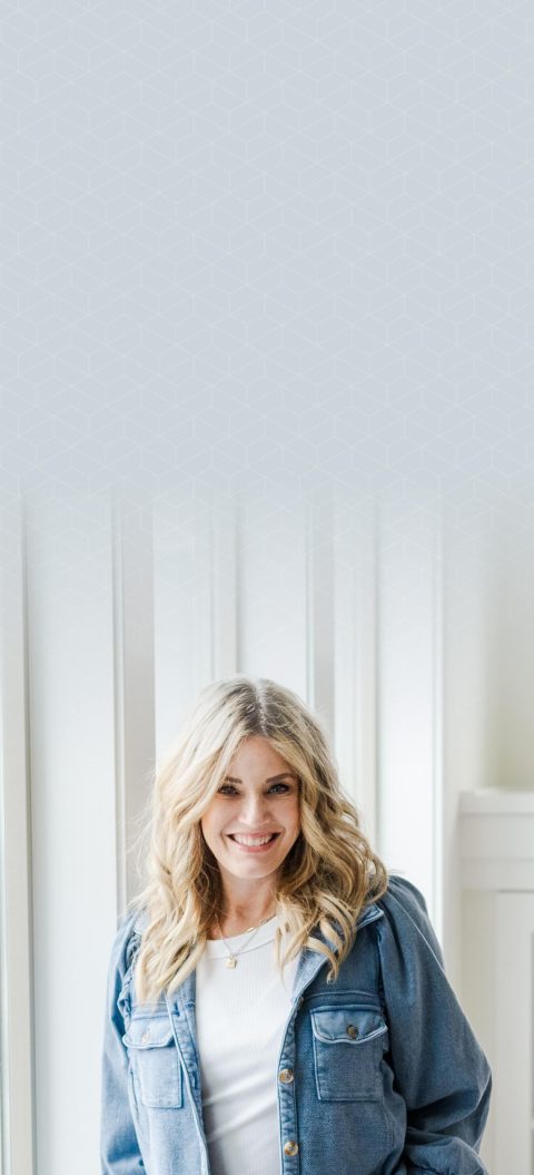 A woman with blonde hair wearing a blue shirt and white top stands indoors, smiling in front of white vertical paneling.