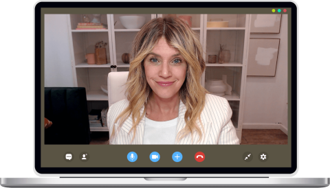 A woman with blonde hair in a white blazer sits in front of shelves, smiling during a video call on a laptop screen.