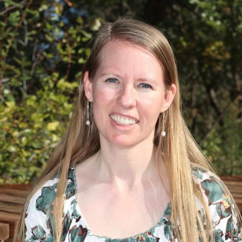 A woman with long blonde hair and pearl earrings is smiling outdoors, sitting in front of green foliage and wooden boards, wearing a patterned top.