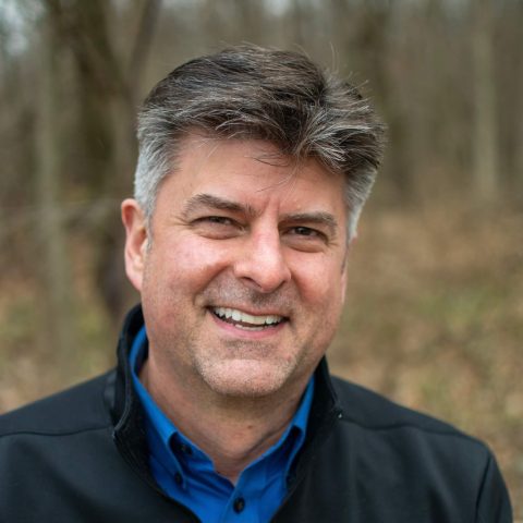 A man with gray hair and a blue collared shirt smiles outdoors with trees and brown foliage in the background.