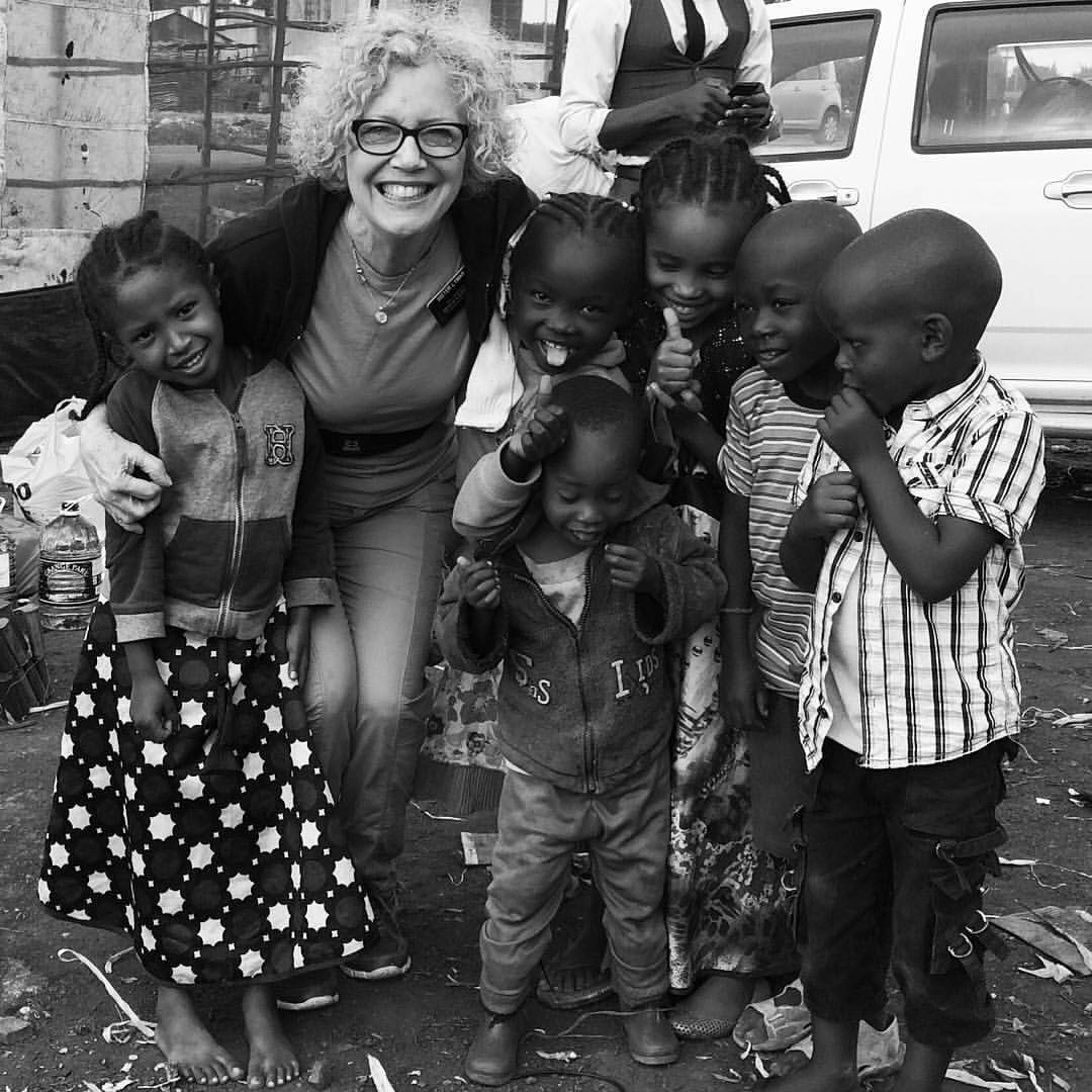 A woman with curly hair smiles and hugs a group of six children, who are standing together outdoors and smiling at the camera.