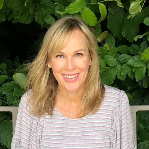 A woman with blonde hair and a striped shirt smiles in front of dense green foliage.