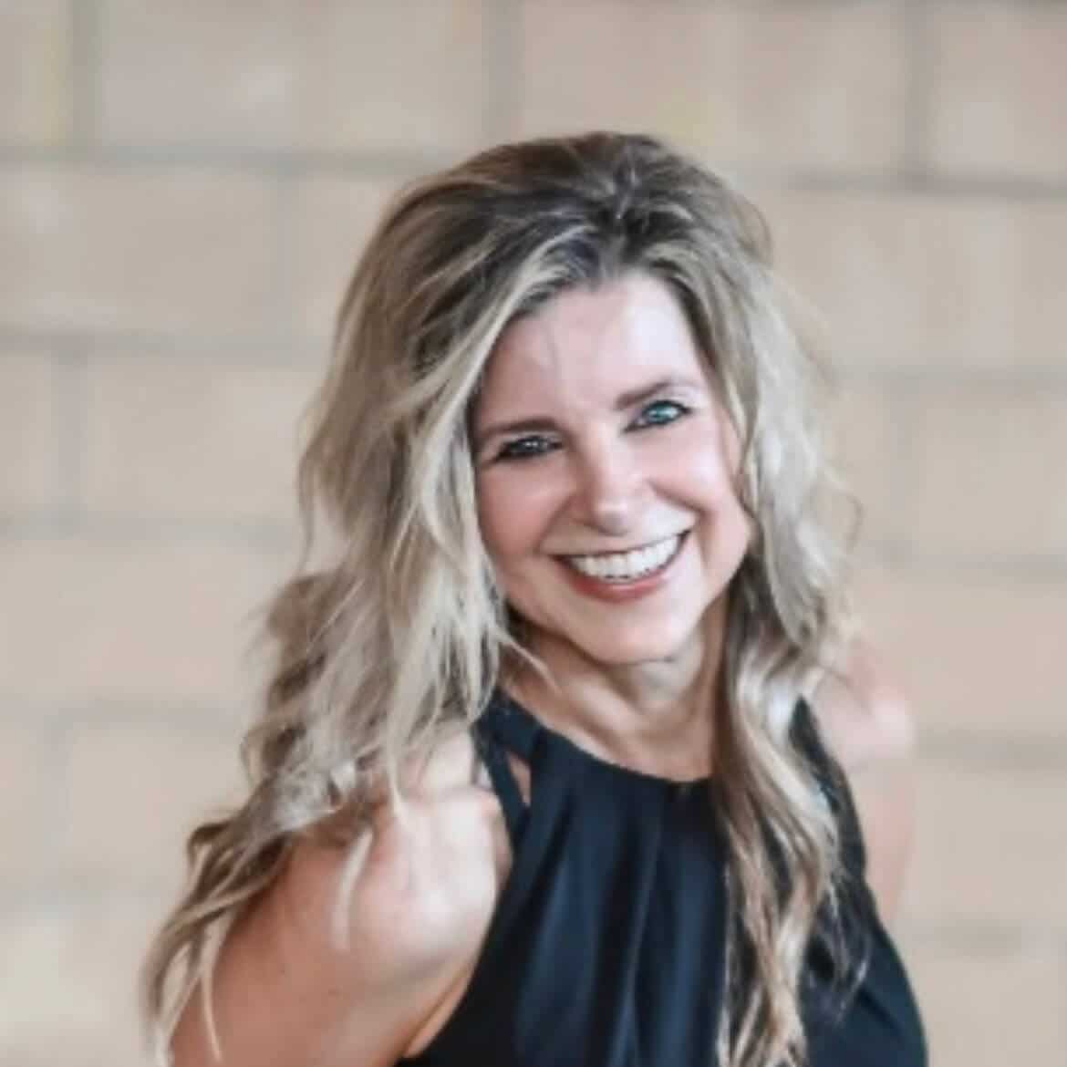 A woman with long, wavy blonde hair and a sleeveless black top smiles at the camera in front of a blurred brick wall background.