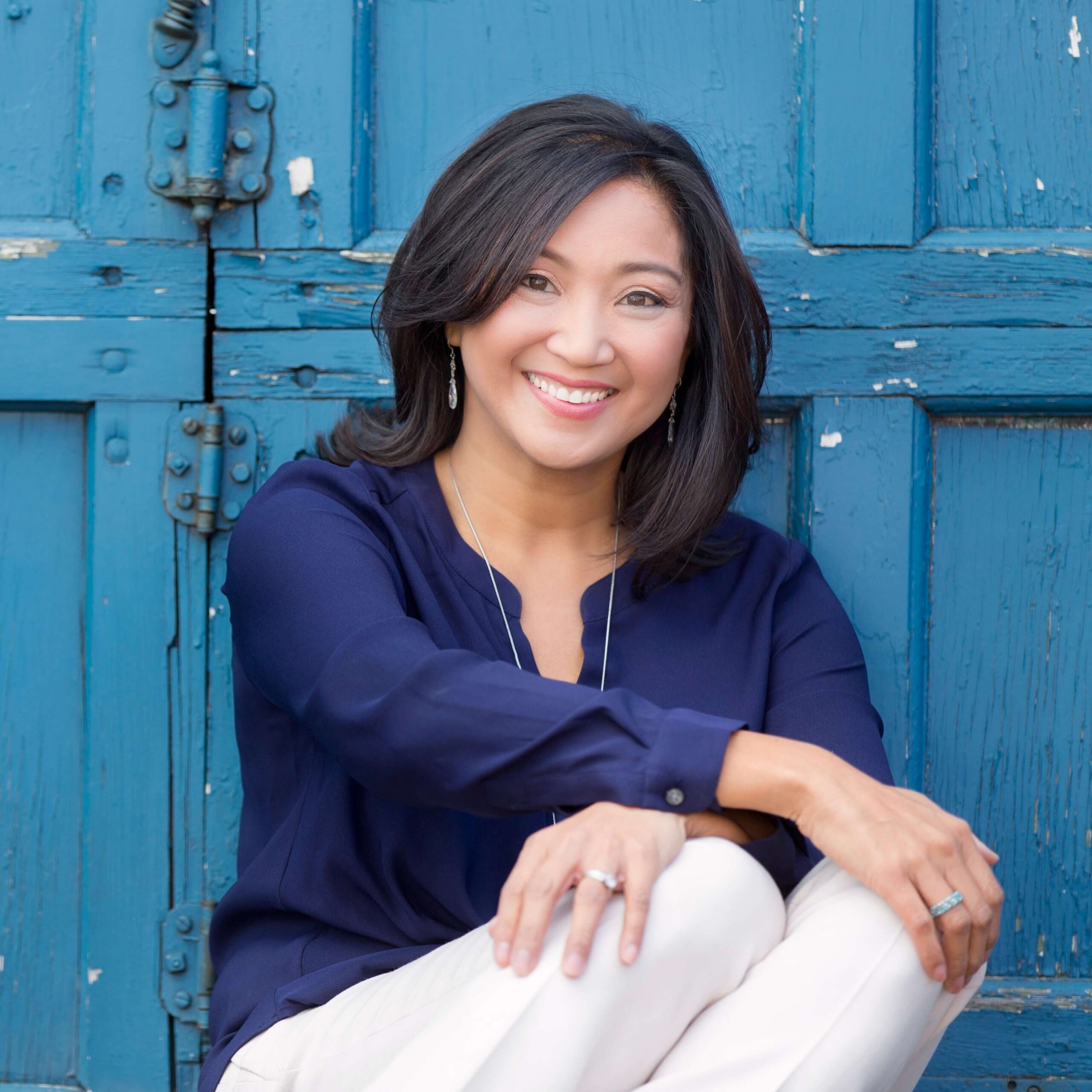 Woman with shoulder-length dark hair, wearing a navy blouse and light pants, sits smiling in front of a weathered blue wooden door.