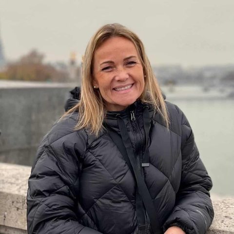 A woman wearing a black puffer jacket stands outdoors by a stone railing, smiling, with a foggy cityscape in the background.