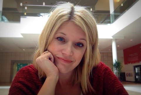 A woman with blonde hair and blue eyes, wearing a red sweater, rests her chin on her hand while sitting indoors in a bright, modern building.
