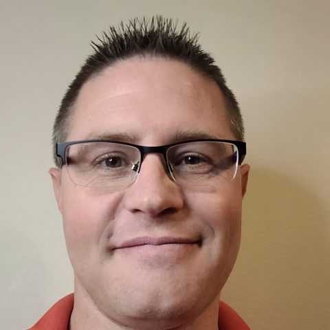 A man with short brown hair, glasses, and a red collared shirt smiles slightly while facing the camera against a plain beige background.