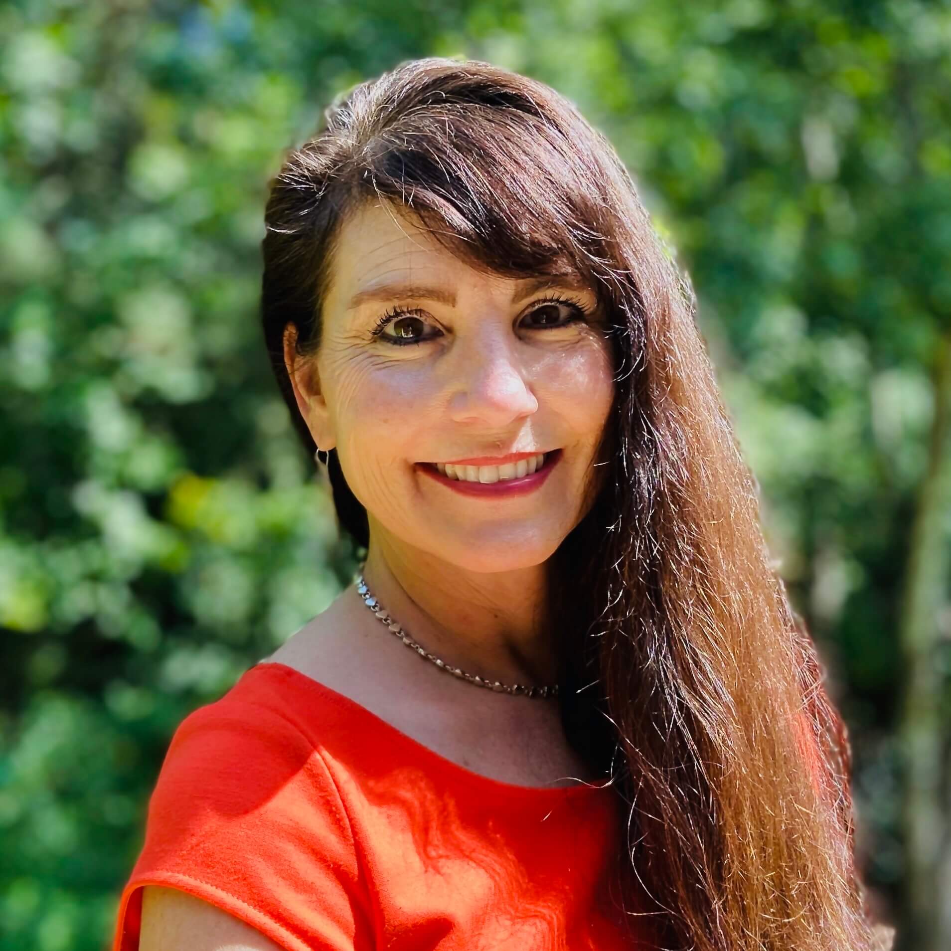 A woman with long brown hair, wearing a red top and necklace, smiles at the camera outdoors with greenery in the background.