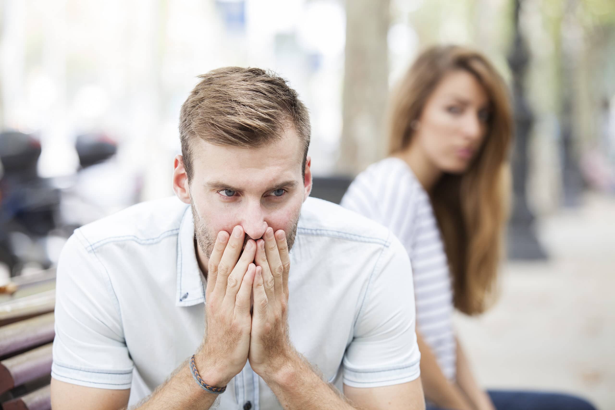 A man sits on a bench looking worried with his hands covering his mouth, while a woman sits behind him, slightly out of focus, appearing concerned.