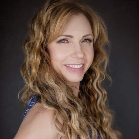 A woman with long wavy blonde hair and a blue patterned top smiles at the camera against a dark background.