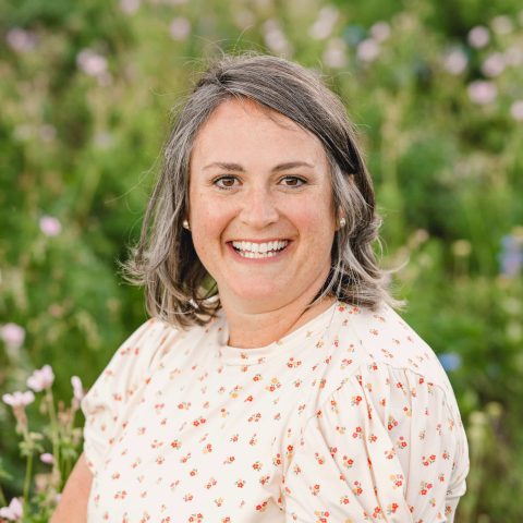 Woman with shoulder-length gray hair wearing a cream-colored, patterned blouse, smiling outdoors in front of a blurred green and floral background.
