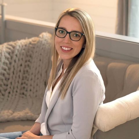 A woman with straight blonde hair and glasses sits on a light-colored sofa, smiling at the camera. She is wearing a light blazer and white top.