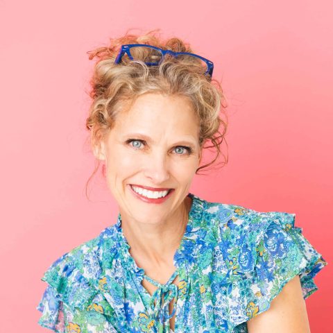 Woman with curly blonde hair, wearing blue glasses on her head and a blue floral blouse, smiling in front of a pink background.
