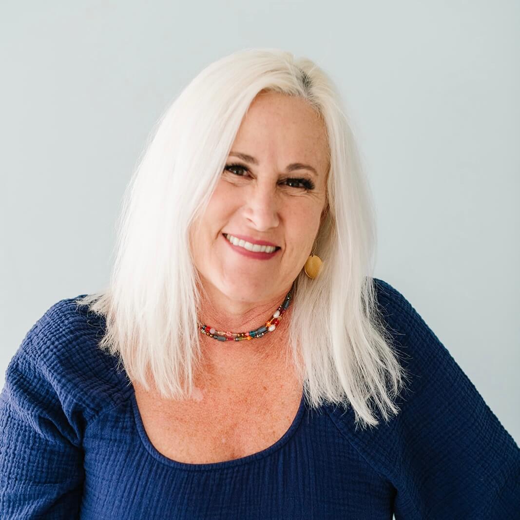 A woman with long white hair wearing a navy blue top and a beaded necklace smiles against a light blue background.