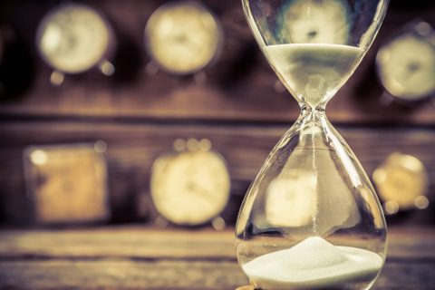 A close-up of an hourglass with sand flowing, with several blurred analog clocks in the background on a wooden surface.