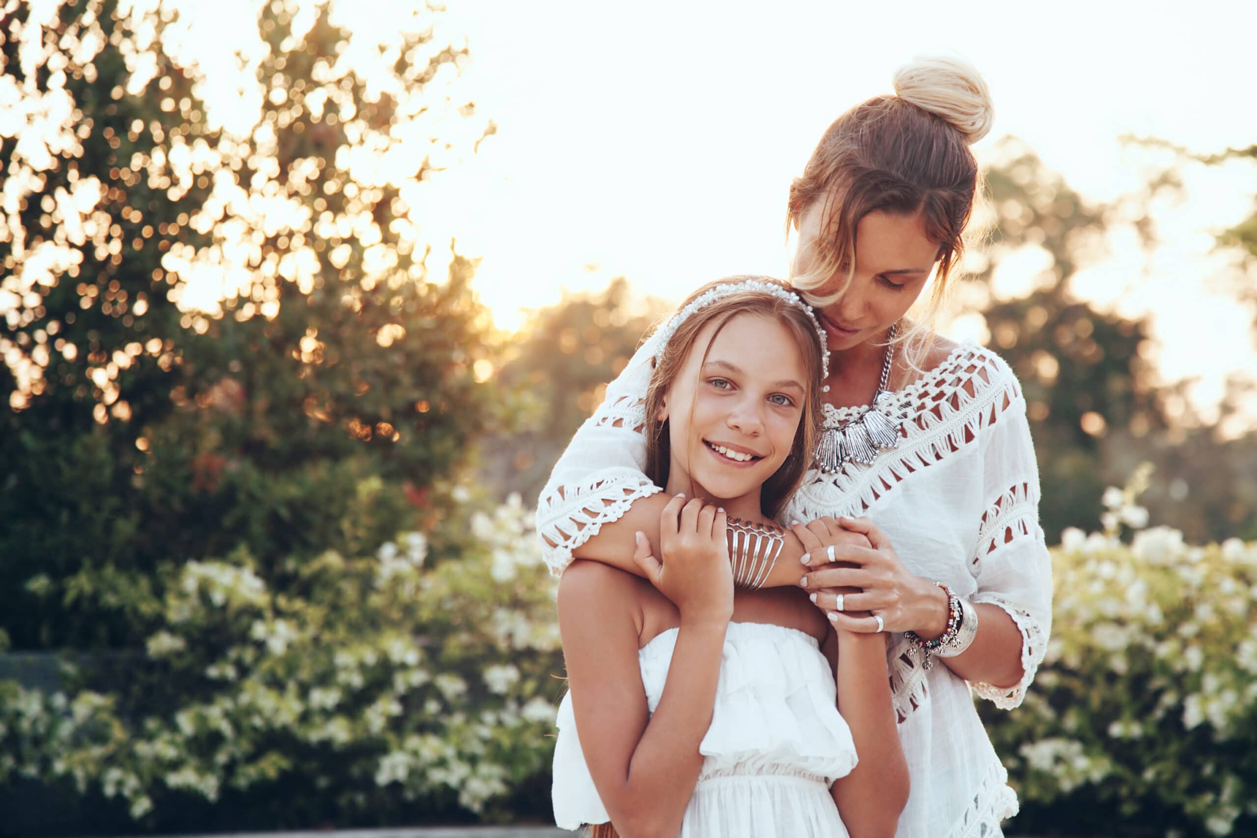 A woman embraces a smiling young girl outdoors, both wearing white clothing, with greenery and sunlight in the background.