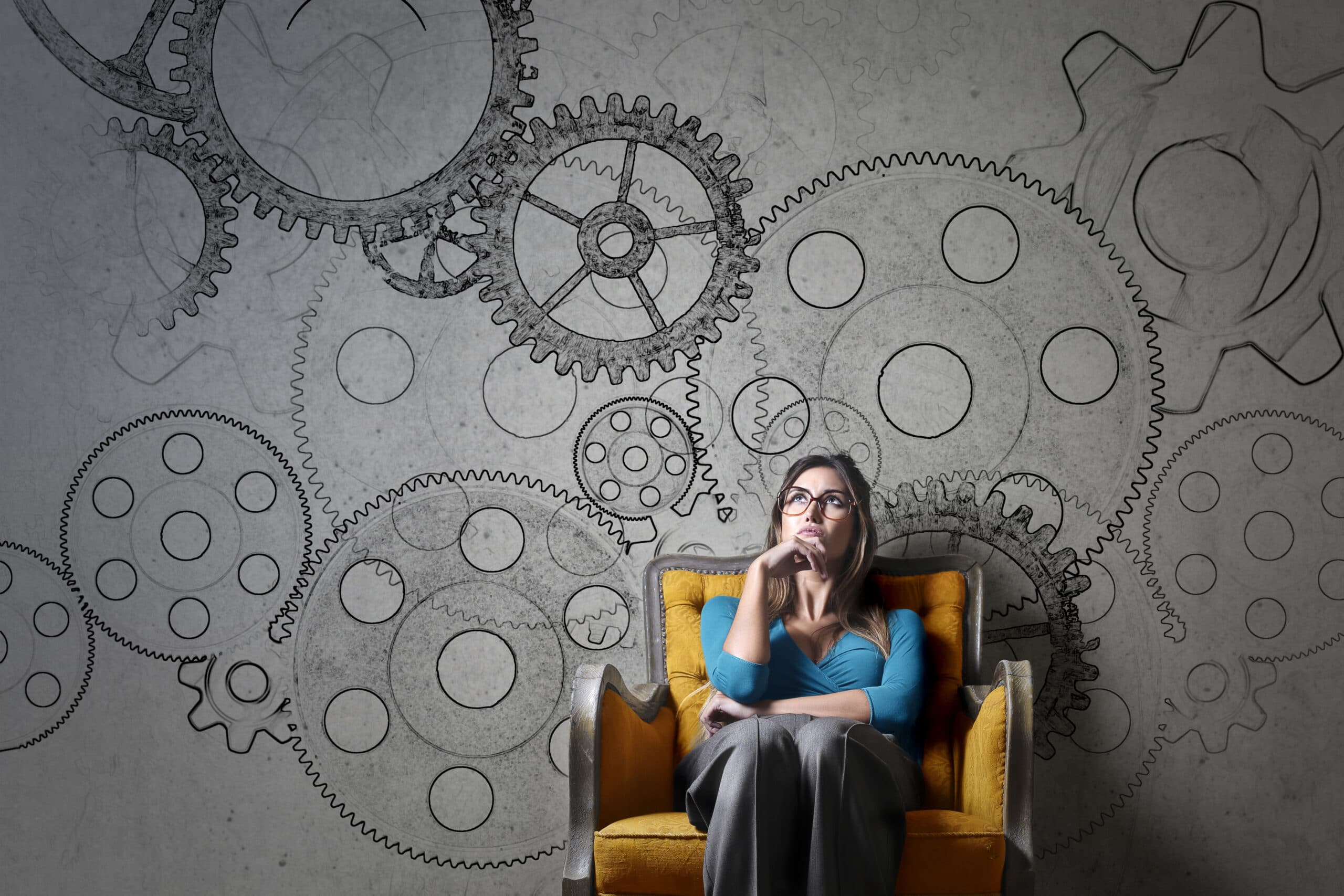 A woman sits in a yellow armchair, appearing thoughtful, with large illustrated gears and cogs drawn on the wall behind her.