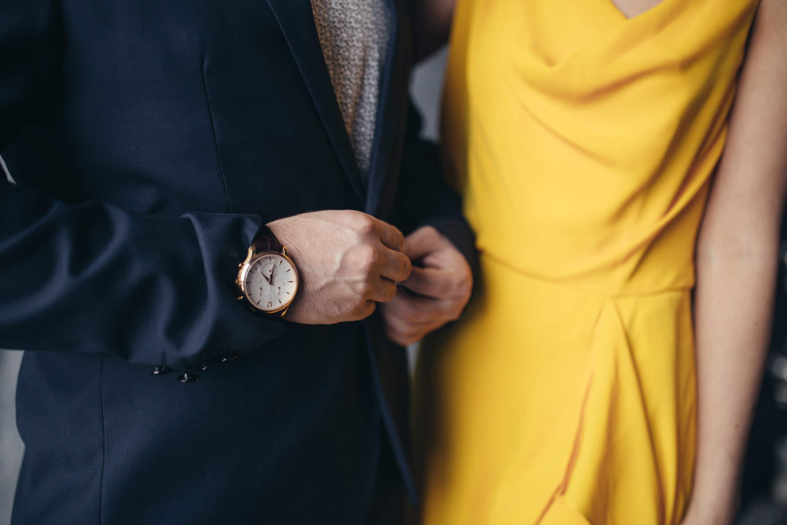 A person in a dark suit adjusts their jacket next to another person wearing a yellow dress. The suited person is wearing a wristwatch.