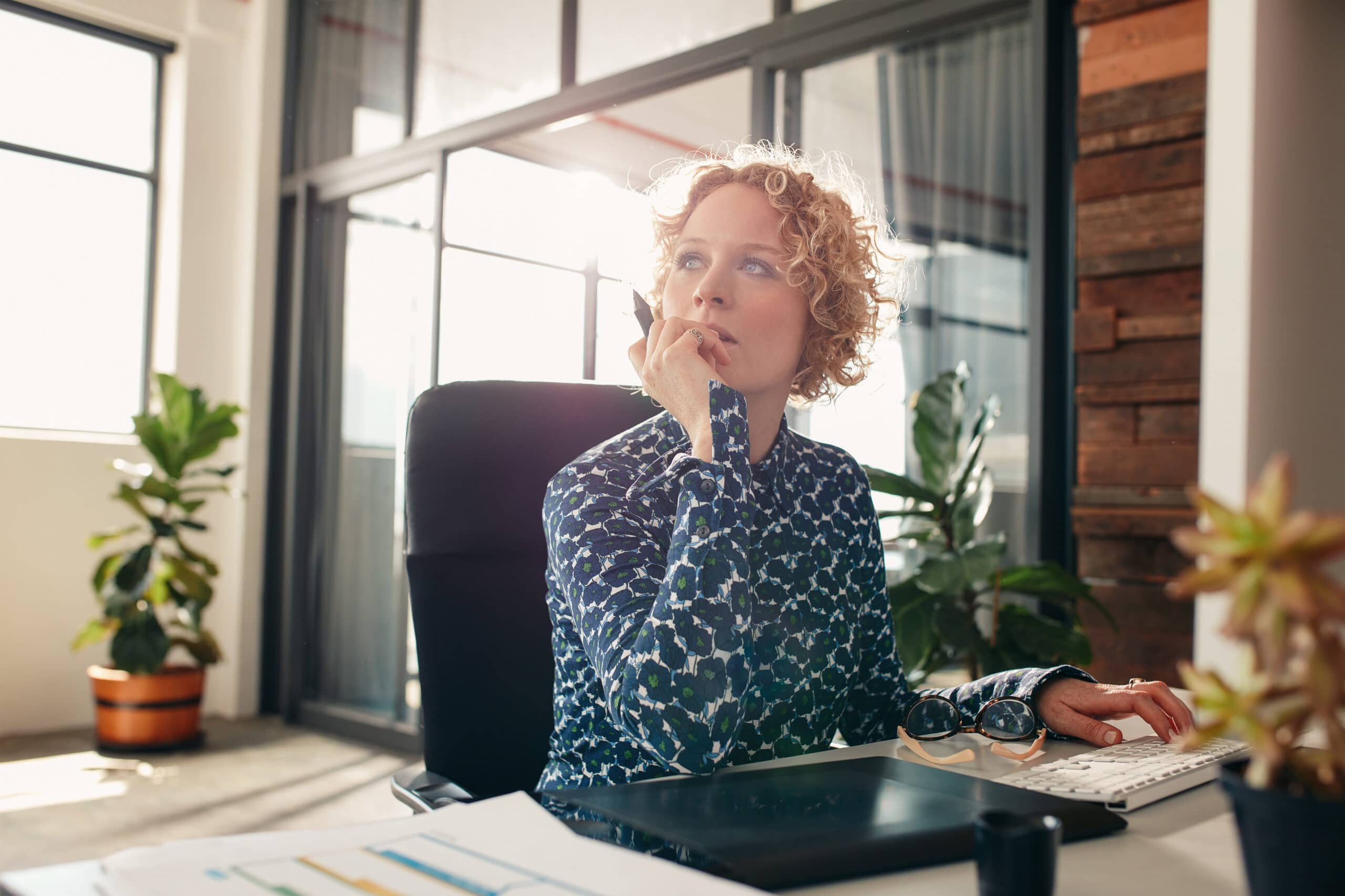 A person with curly hair sits at a desk in a modern office, holding a pen and looking thoughtful, with plants and sunlight in the background.