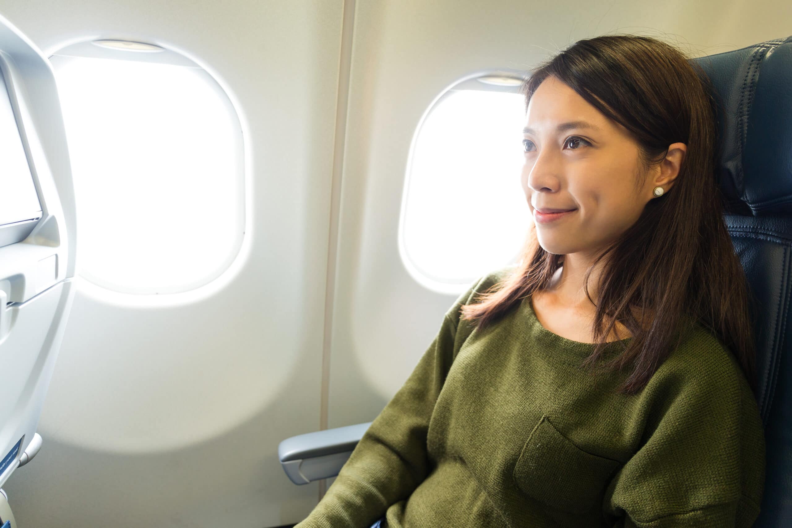 A woman with long hair and a green sweater sits by an airplane window, facing forward with a slight smile.