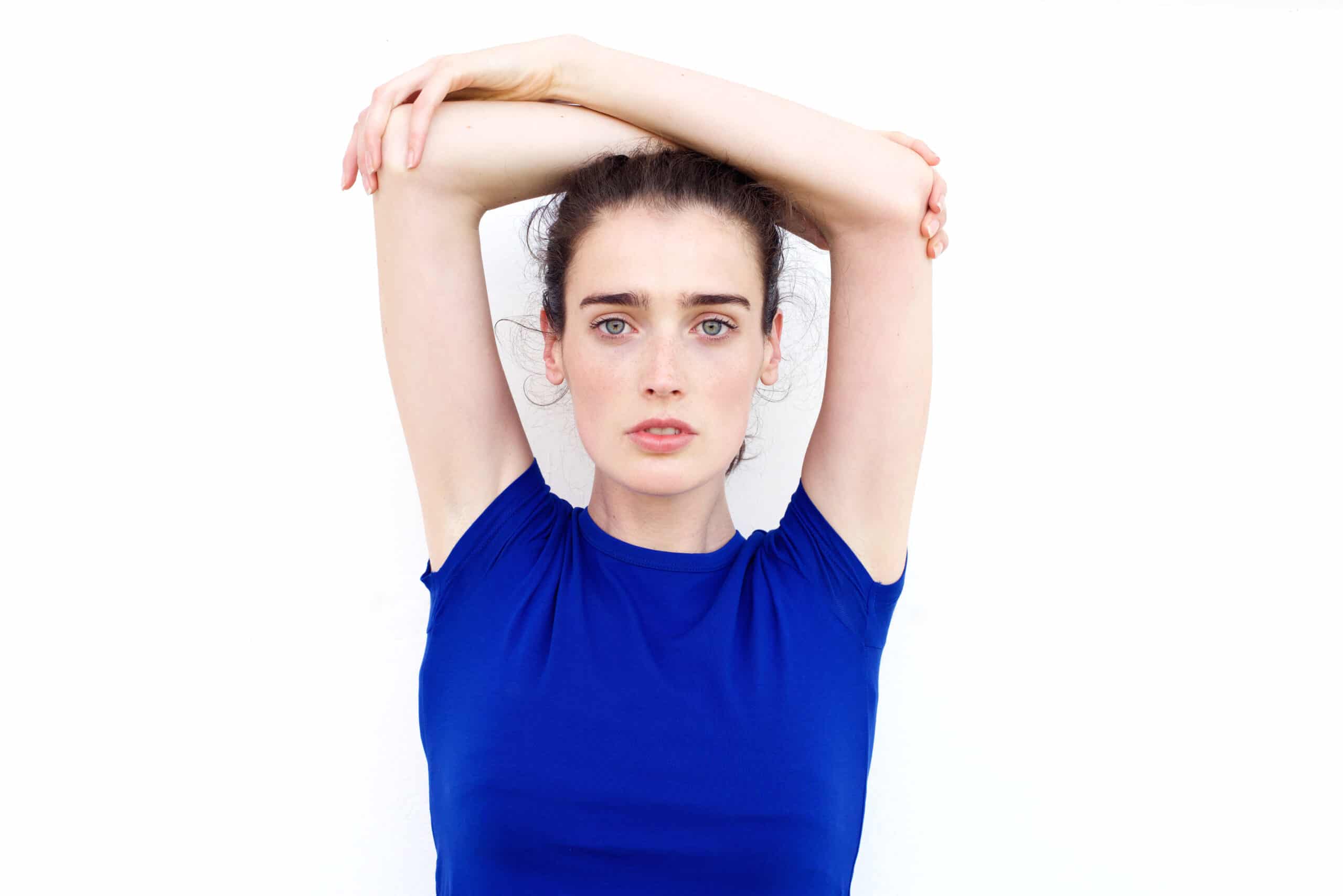 A person wearing a blue shirt stands against a white background with arms raised and crossed above their head, looking directly at the camera.