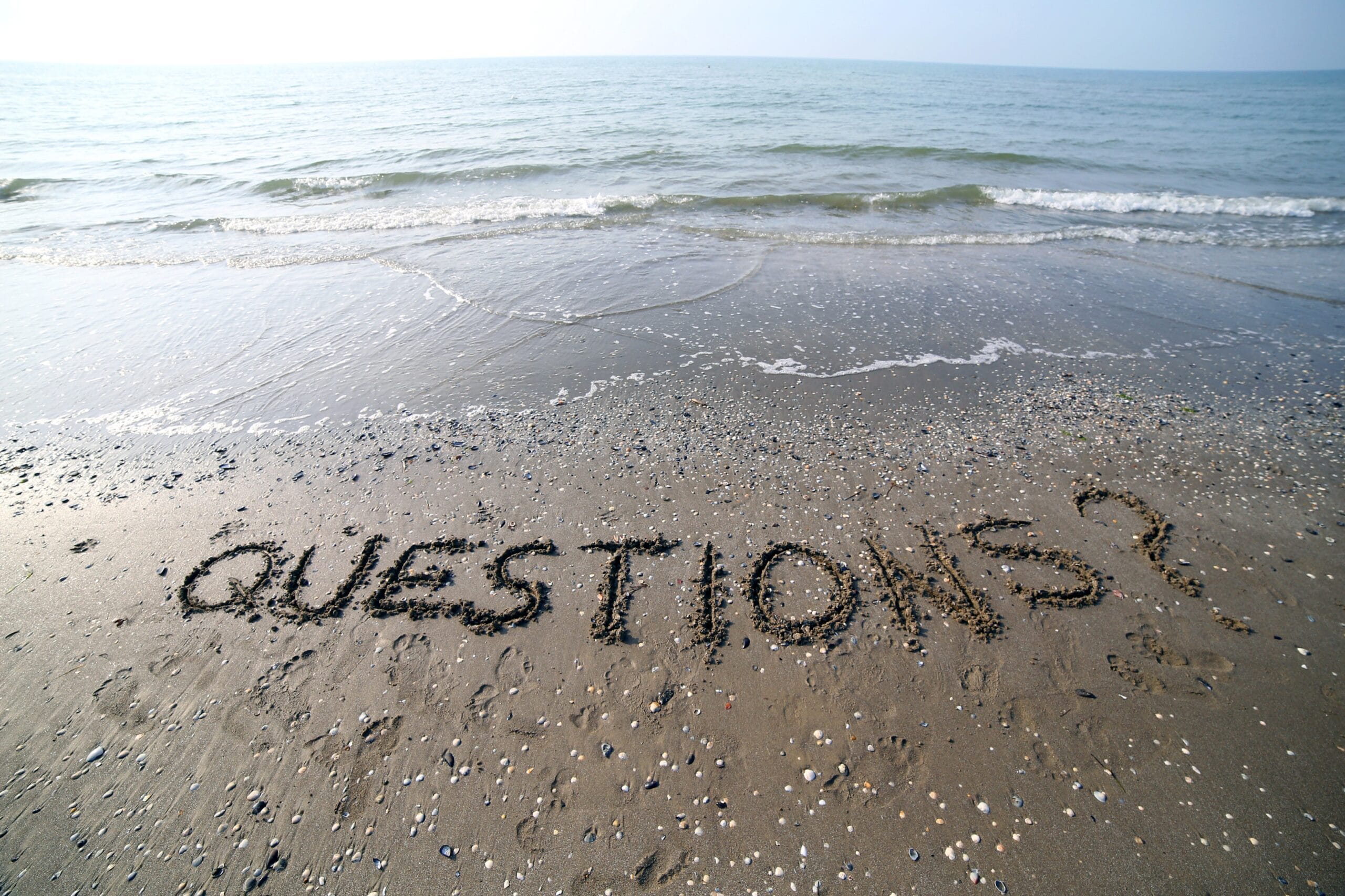 The word "QUESTIONS?" is written in the sand near the shoreline, with gentle waves and the sea visible in the background.
