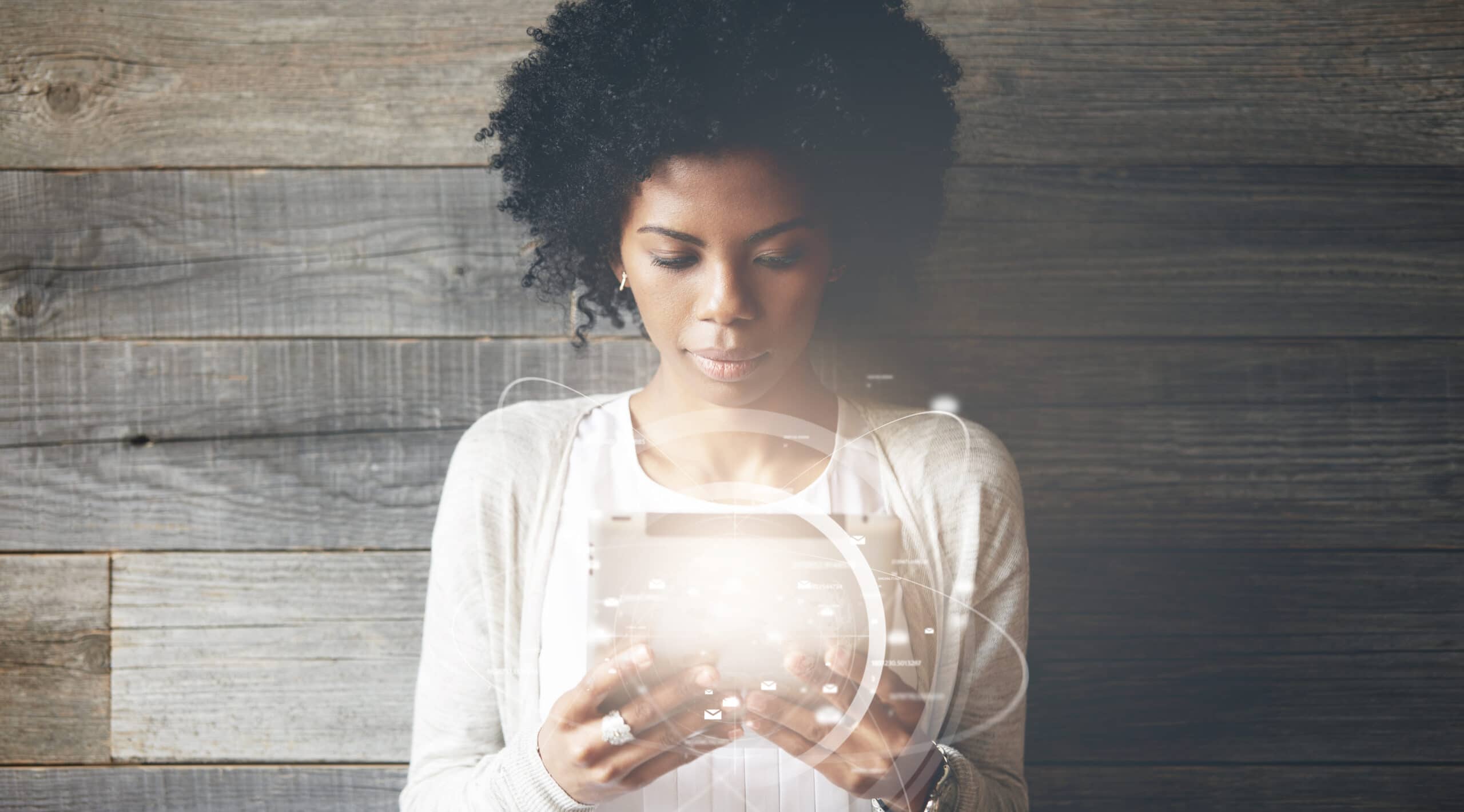 A woman with curly hair stands against a wooden wall, looking down at a glowing tablet with digital interface graphics overlayed.