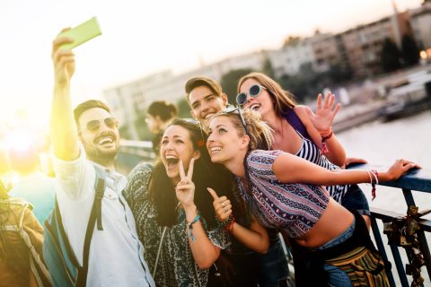 A group of five young adults stand on a bridge, smiling and posing for a selfie with a smartphone near sunset.