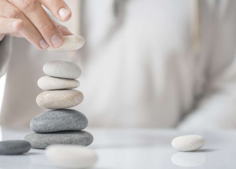 A hand stacking smooth, flat stones into a balanced tower on a reflective surface, with another stone lying nearby.