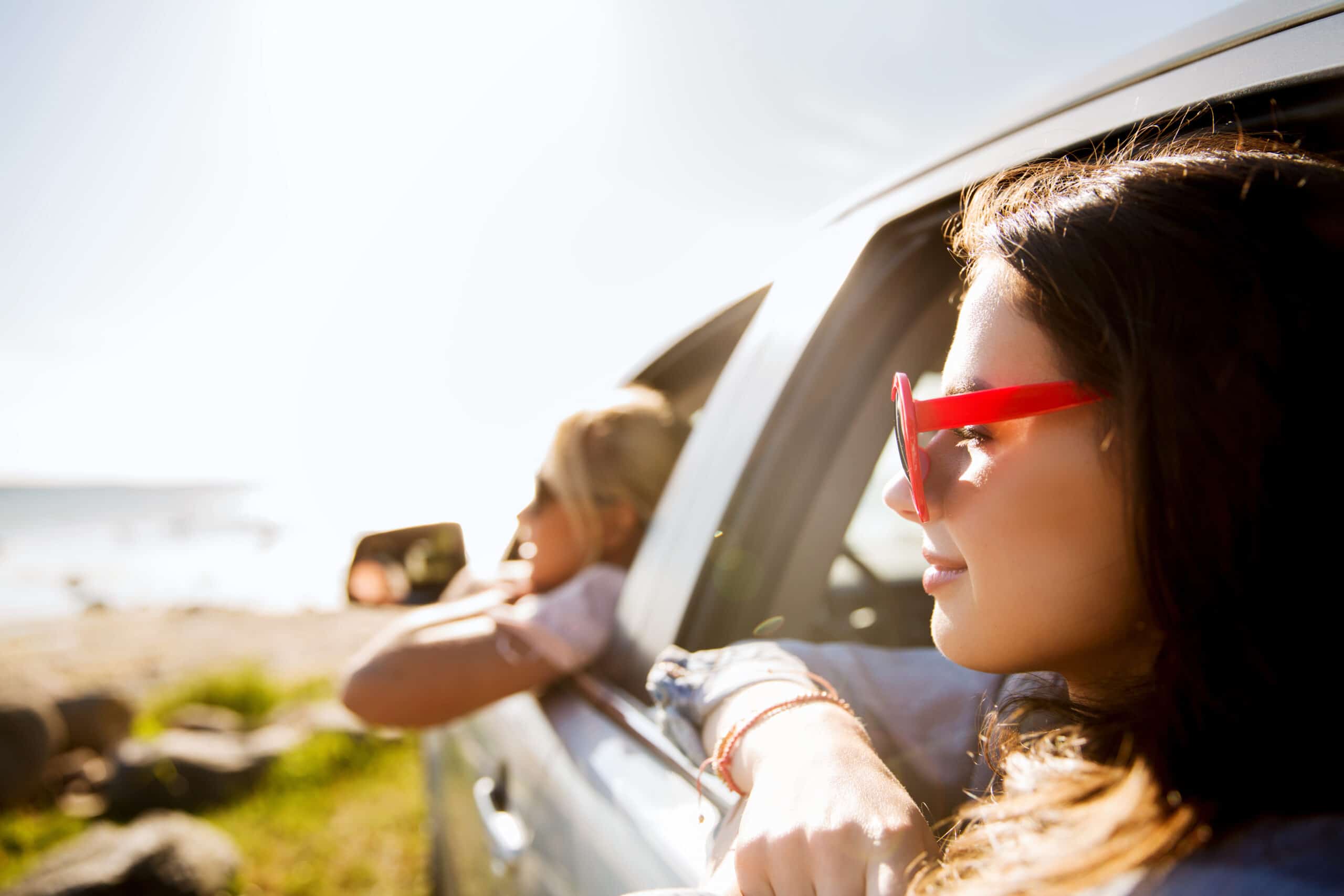 Two people lean out of a car window, looking toward the beach; one wears red sunglasses. The scene is sunny with a blurred shoreline in the background.