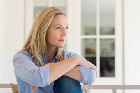 Woman with blonde hair sits indoors, resting her arms on her knees and looking thoughtfully into the distance. She wears a light blue shirt and jeans.