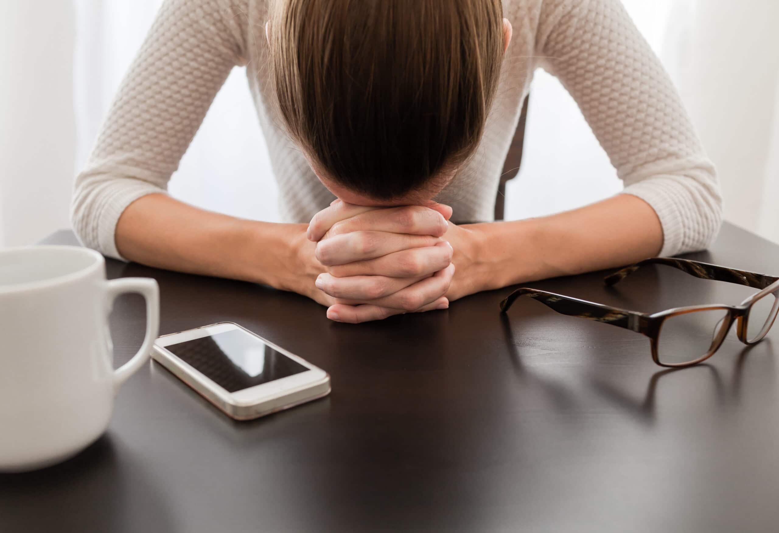 A person sits at a table with their head resting on clasped hands, surrounded by a smartphone, eyeglasses, and a coffee mug.