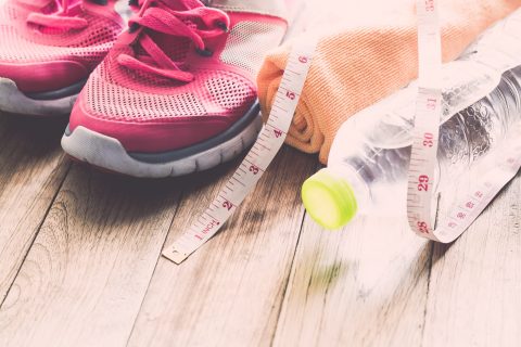 Pink athletic shoes, a measuring tape, an orange towel, and a plastic water bottle arranged on a wooden floor.