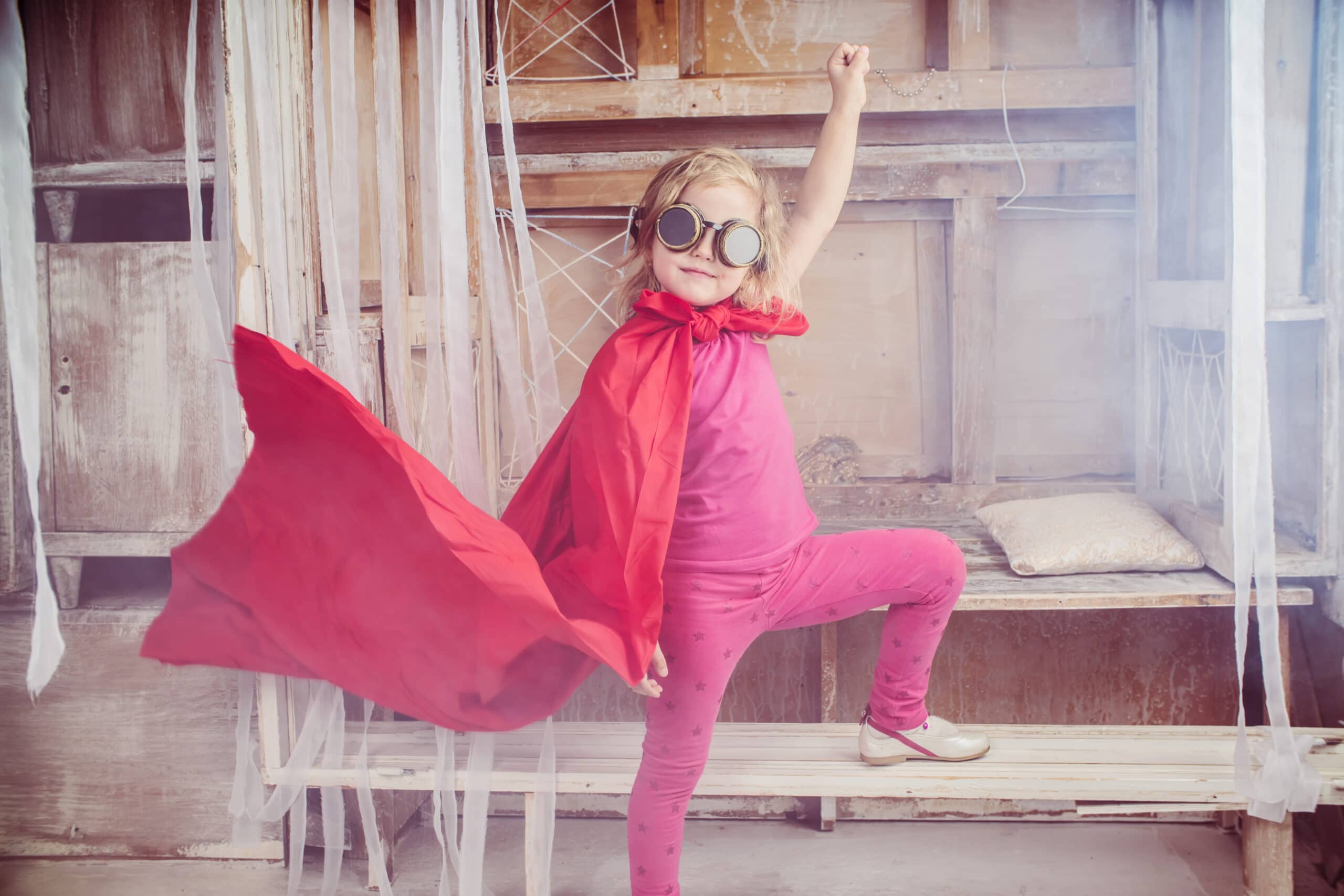 Young child wearing a red cape and goggles poses with one arm raised, standing on a bench in a rustic, decorated room.