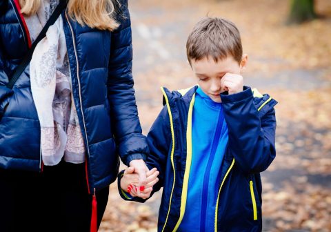 Adult holding hands with a young boy who appears upset, wiping his eyes, while walking outdoors on a path covered with autumn leaves.