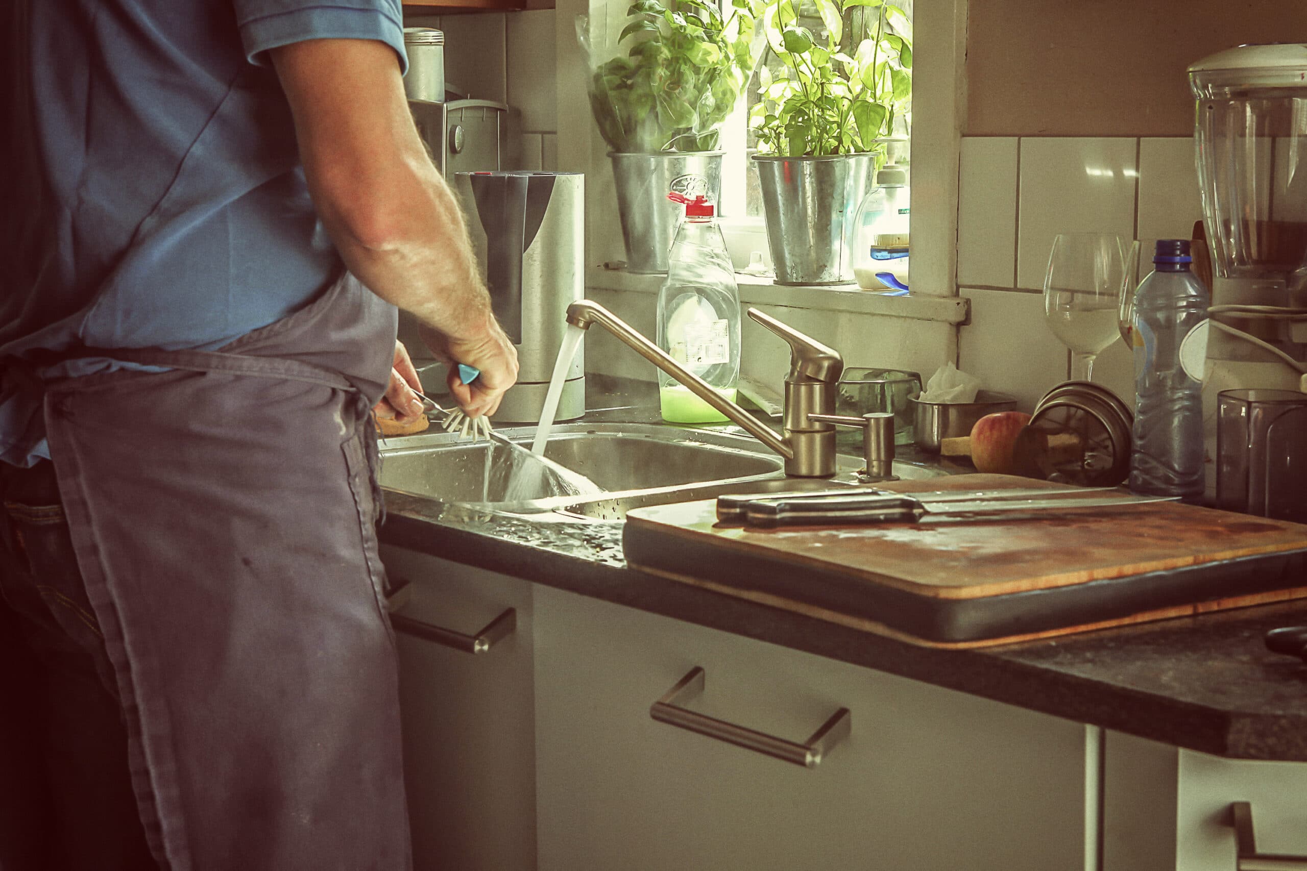 Person wearing an apron washing dishes at a kitchen sink, with a cutting board, knives, and various kitchen items visible on the counter.