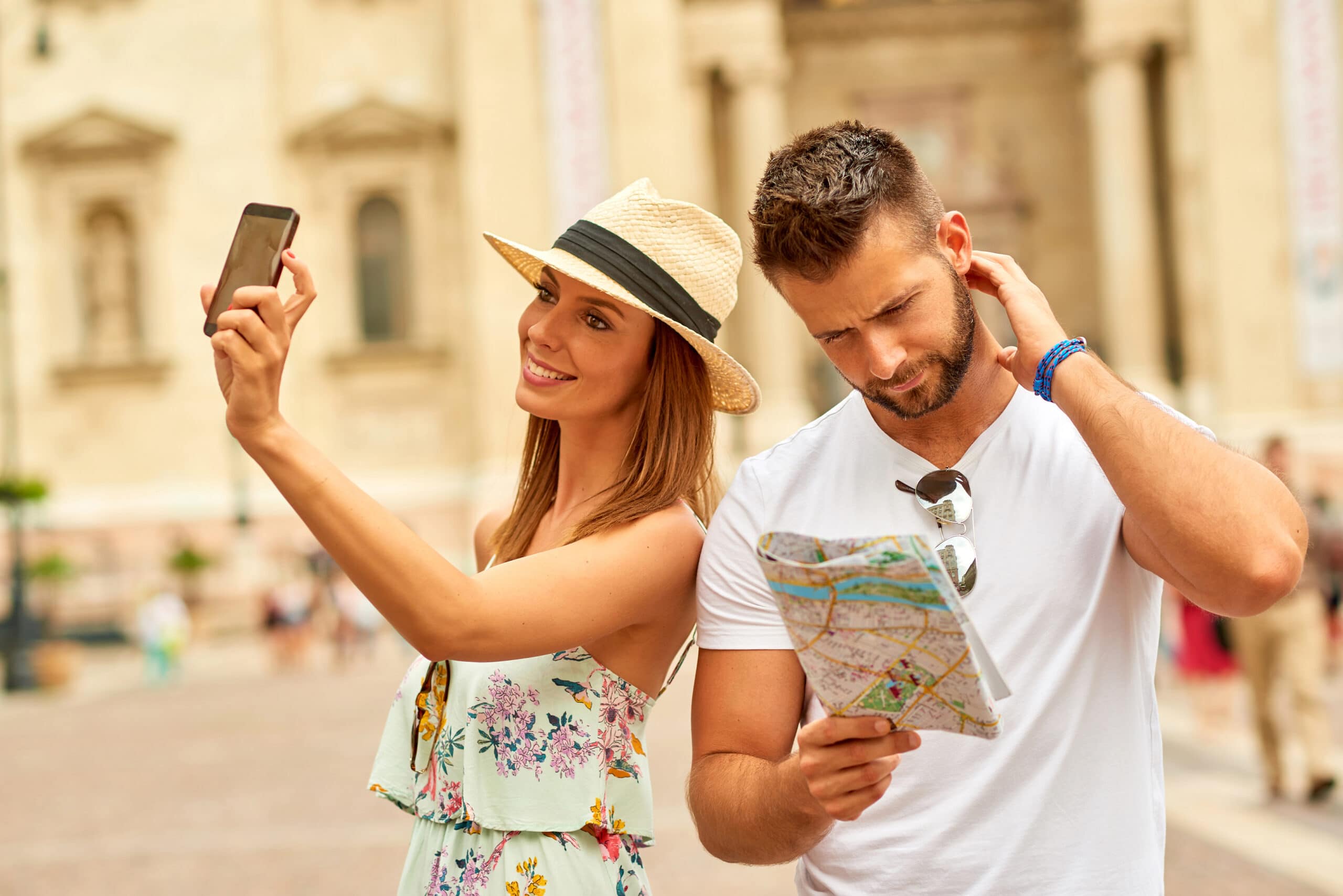 A woman in a hat takes a selfie while a man next to her looks at a map and scratches his head, outdoors in a city setting.