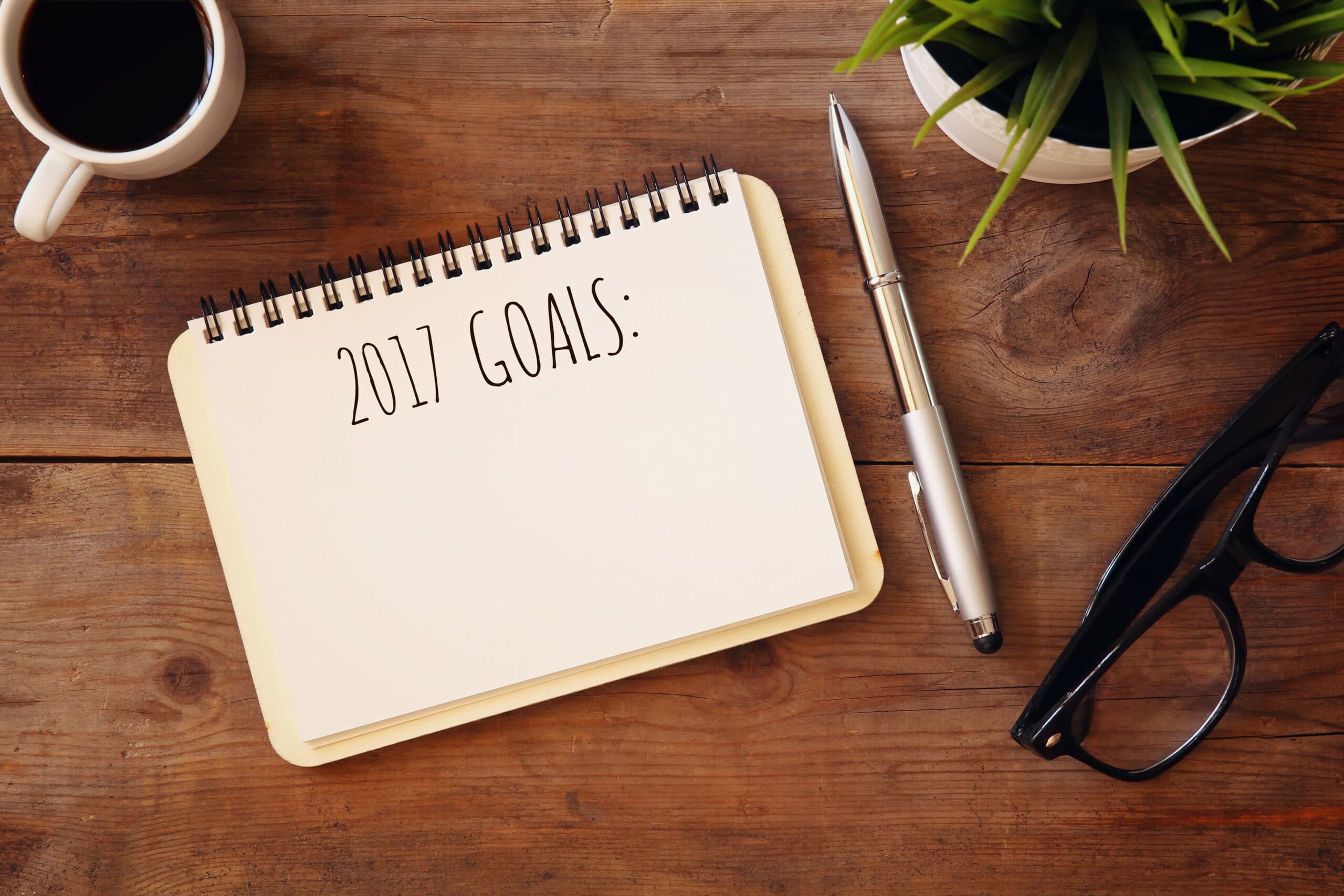 Open notebook titled "2017 Goals:" on a wooden desk with a pen, coffee cup, eyeglasses, and a potted plant nearby.