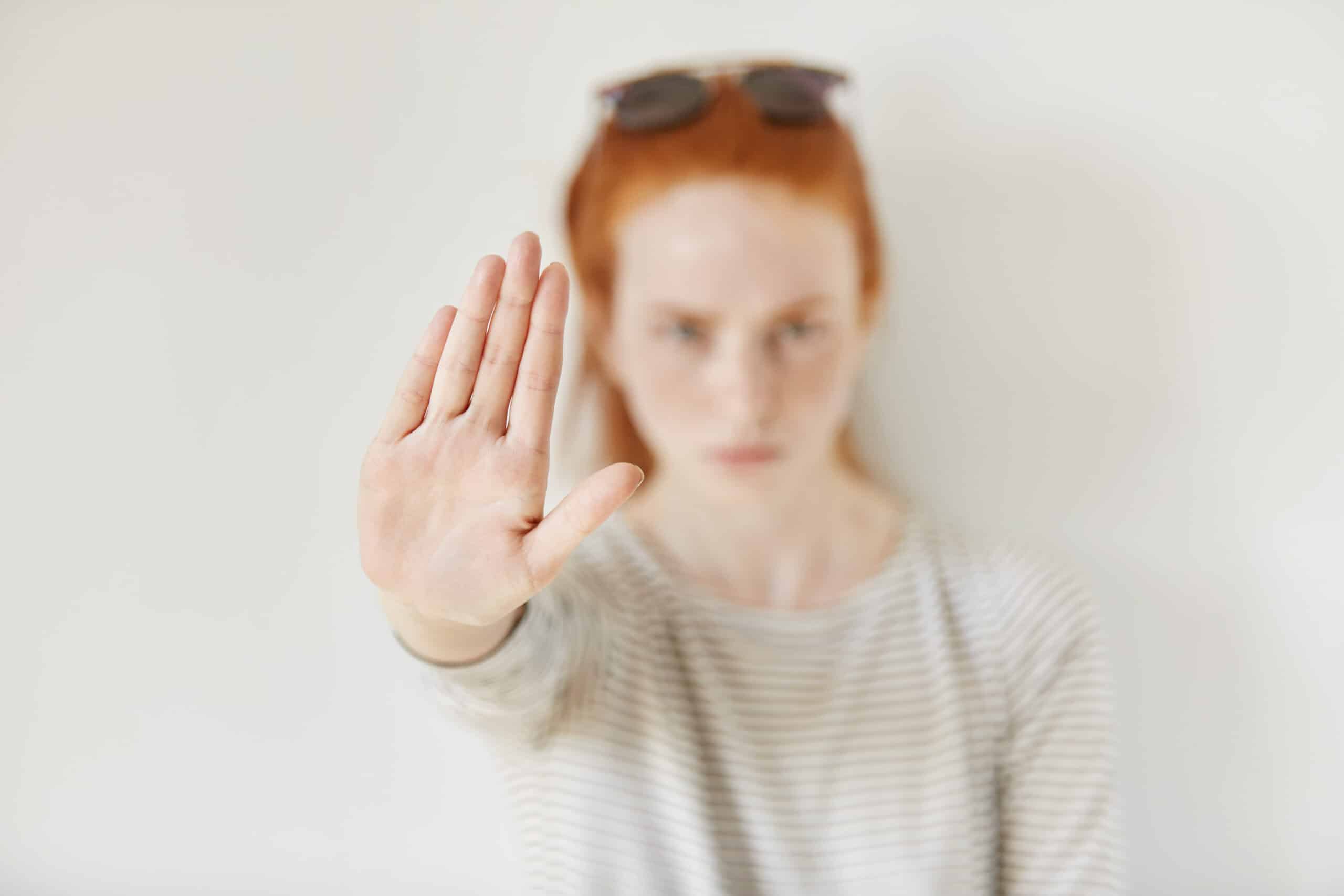 A person with red hair holds up their hand with the palm facing the camera, signaling stop, against a plain white background.