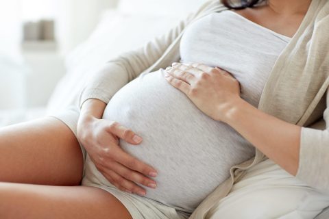 A pregnant woman in light-colored clothes sits on a bed, resting her hands on her bare belly.