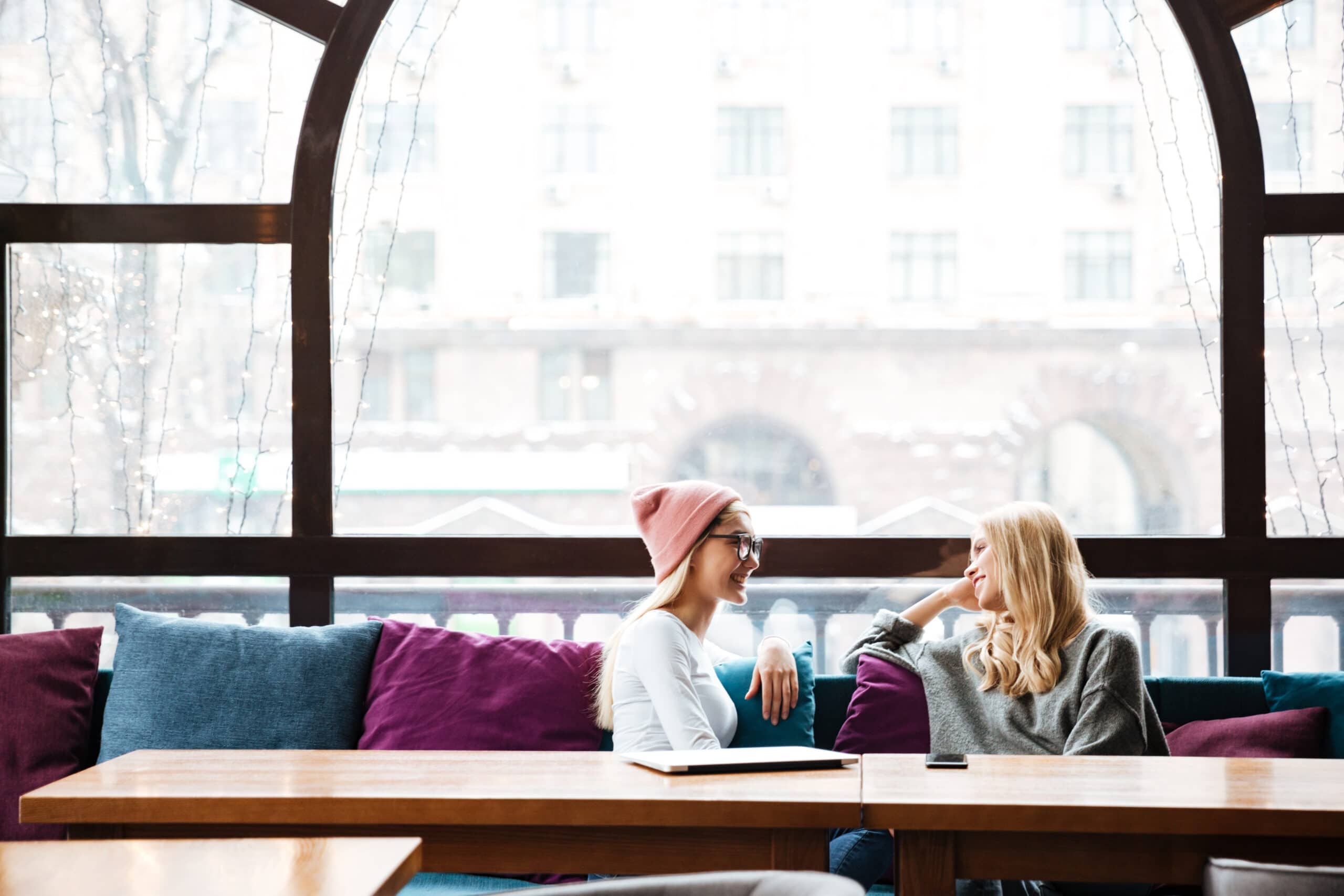 Two women sit on a sofa in a cafe by a large window, facing each other and talking. A smartphone is on the table in front of them.