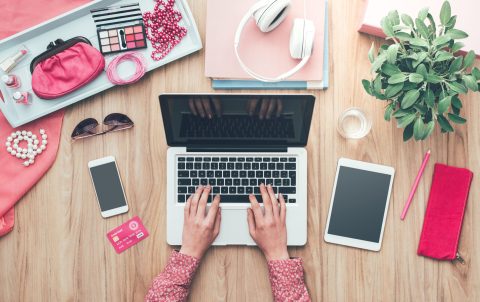 A person types on a laptop at a wooden desk with a smartphone, tablet, credit card, headphones, makeup, accessories, a notebook, and a potted plant.