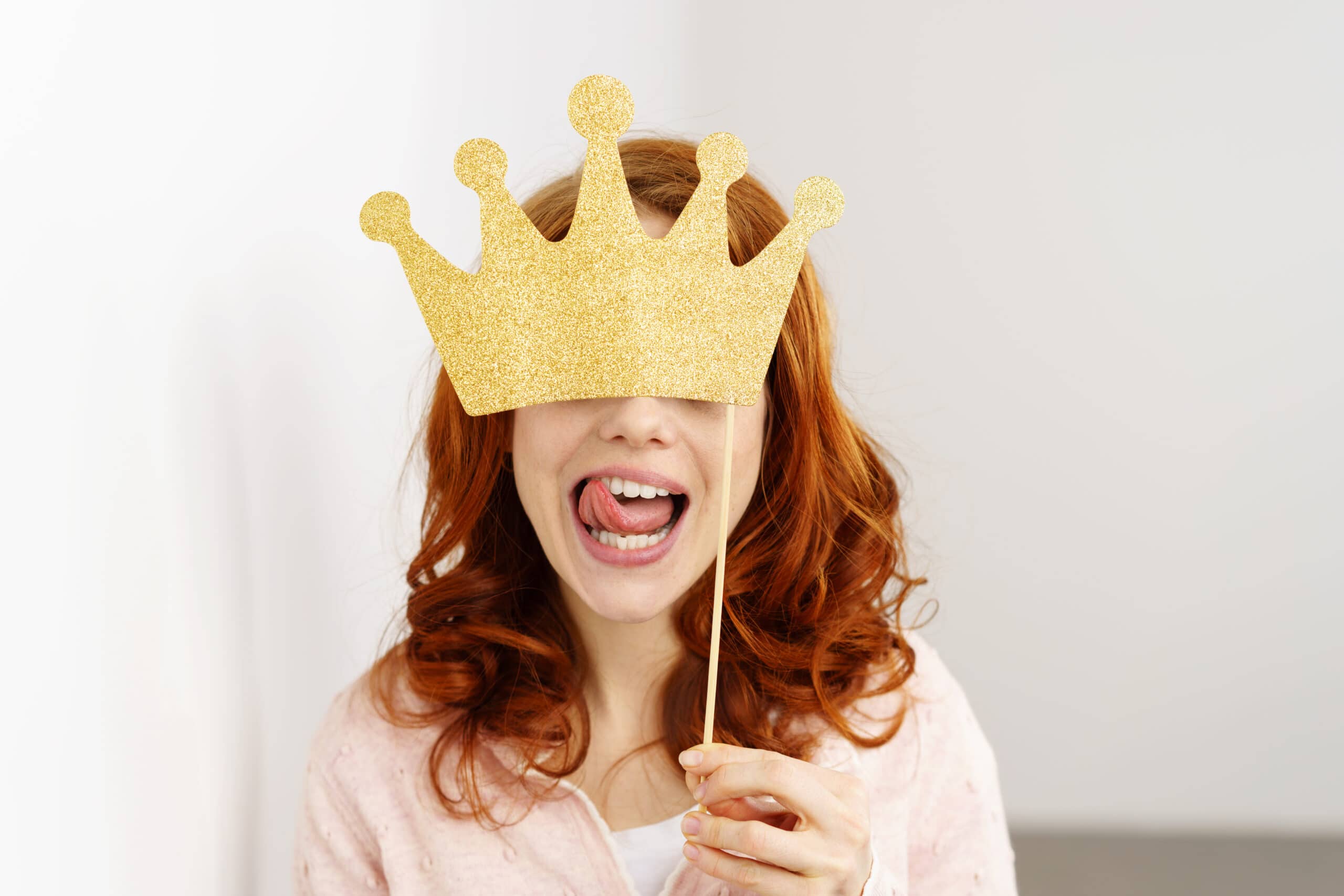 A person with red hair holds a glittery gold paper crown on a stick in front of their face while sticking out their tongue.