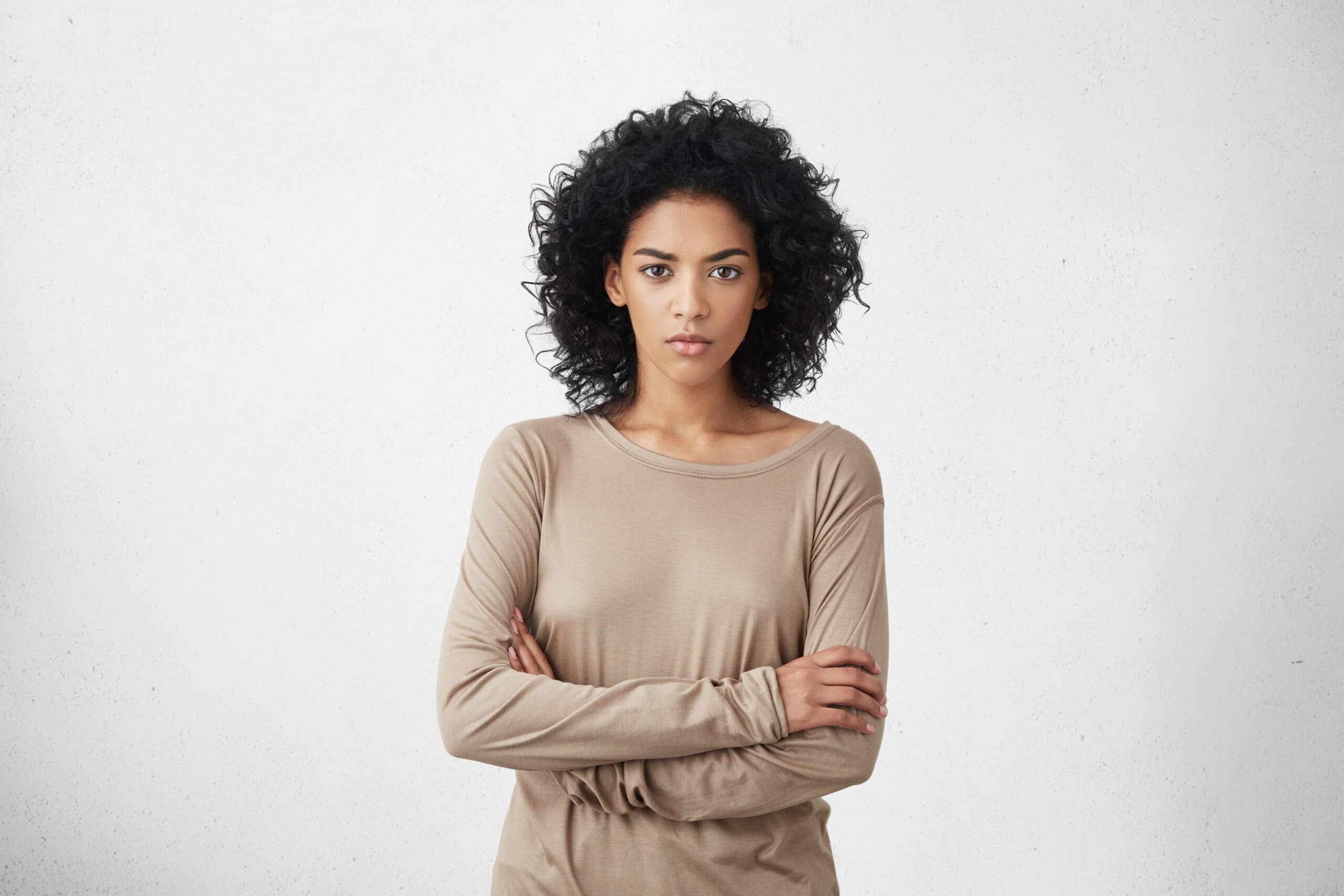 Woman with curly hair wearing a beige long-sleeve shirt stands against a plain white background with her arms crossed, looking straight at the camera.