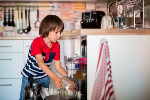 A young boy in a red and blue striped shirt loads dishes into a dishwasher in a modern kitchen.