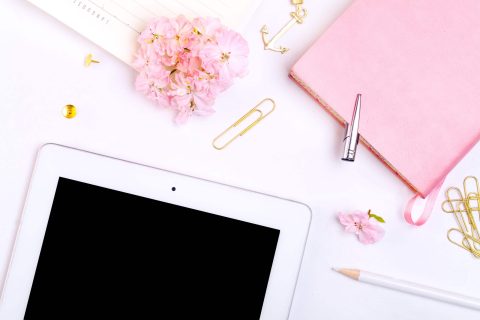 A tablet, pink notebook, pen, pencil, paper clips, flowers, and notepad are arranged on a white desk.