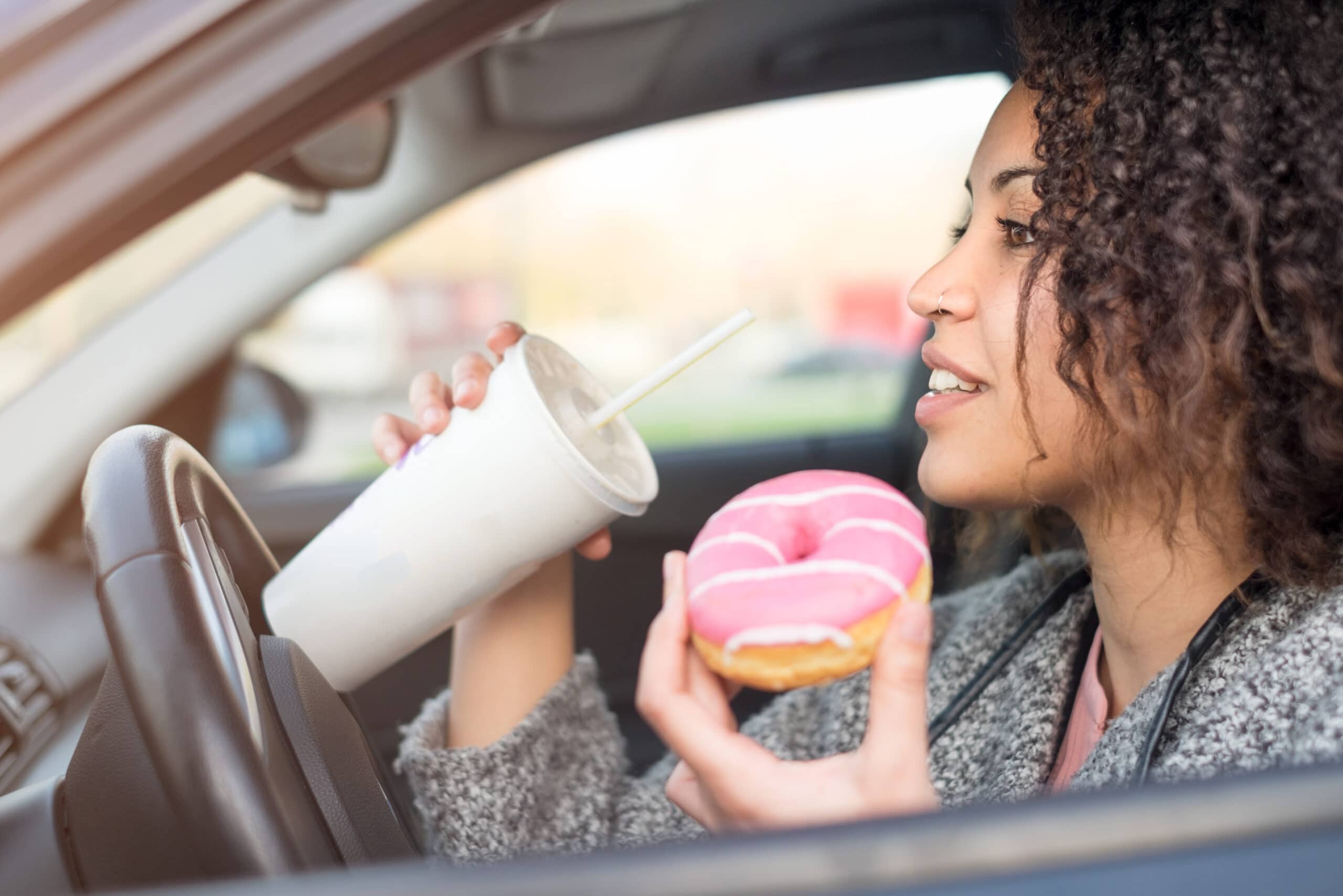 A woman sits in a car, holding a pink frosted donut in one hand and a drink with a straw in the other.