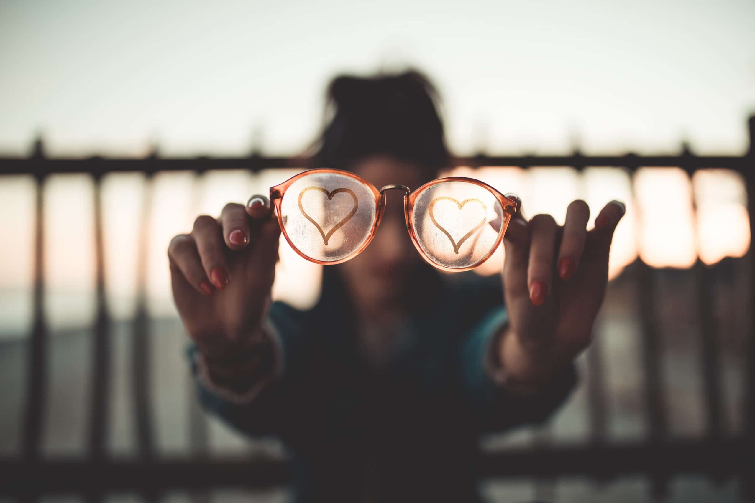 Person holding out glasses with heart shapes drawn on each lens, standing in front of a fence with blurred background.