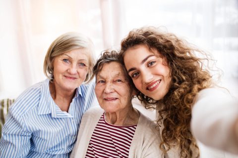 Three women from different generations sit closely together, smiling at the camera as they take a selfie indoors.