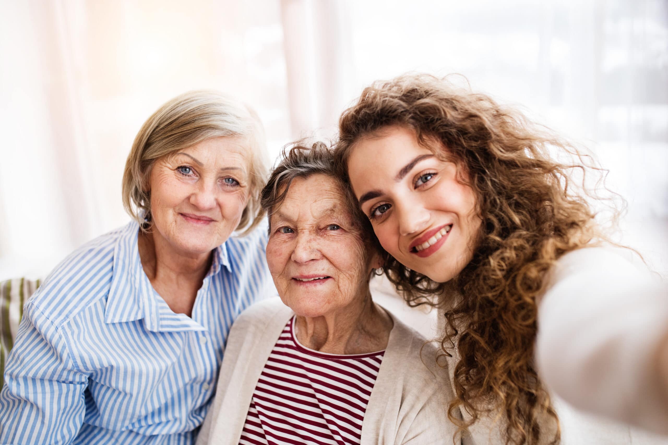 Three women from different generations sit closely together, smiling at the camera as they take a selfie indoors.